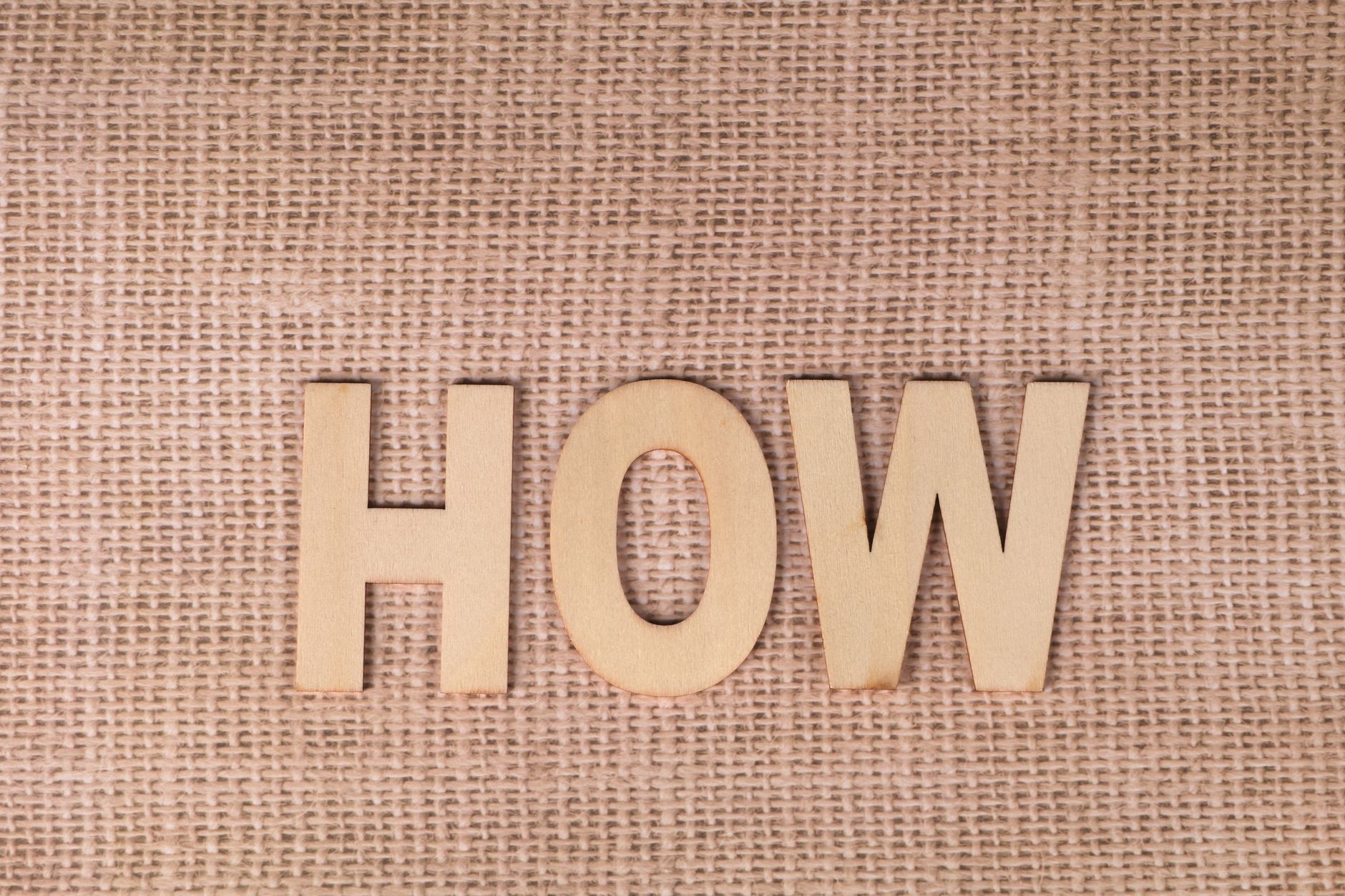 Word 'HOW' formed with wooden letters on textured burlap surface.